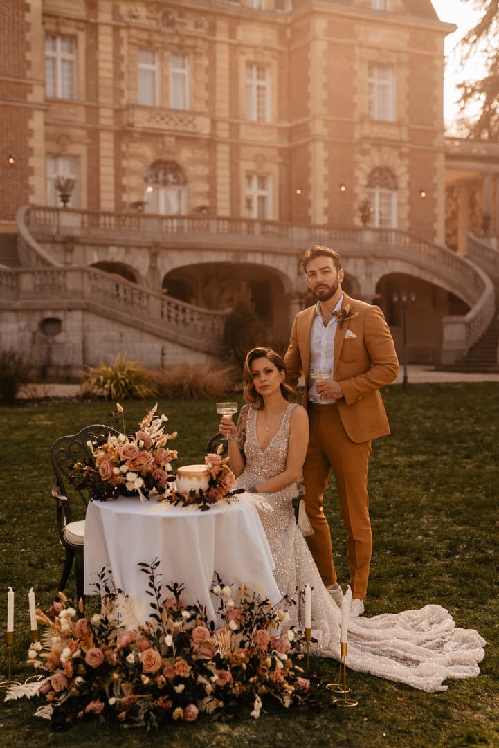 A bride and groom pose with champagne beside a flowered table in a golden-lit garden