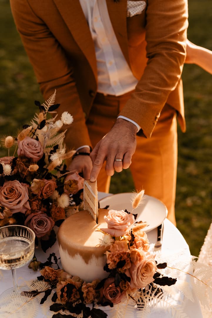 Groom in a rust suit cuts a small wedding cake surrounded by roses at a sunlit garden table