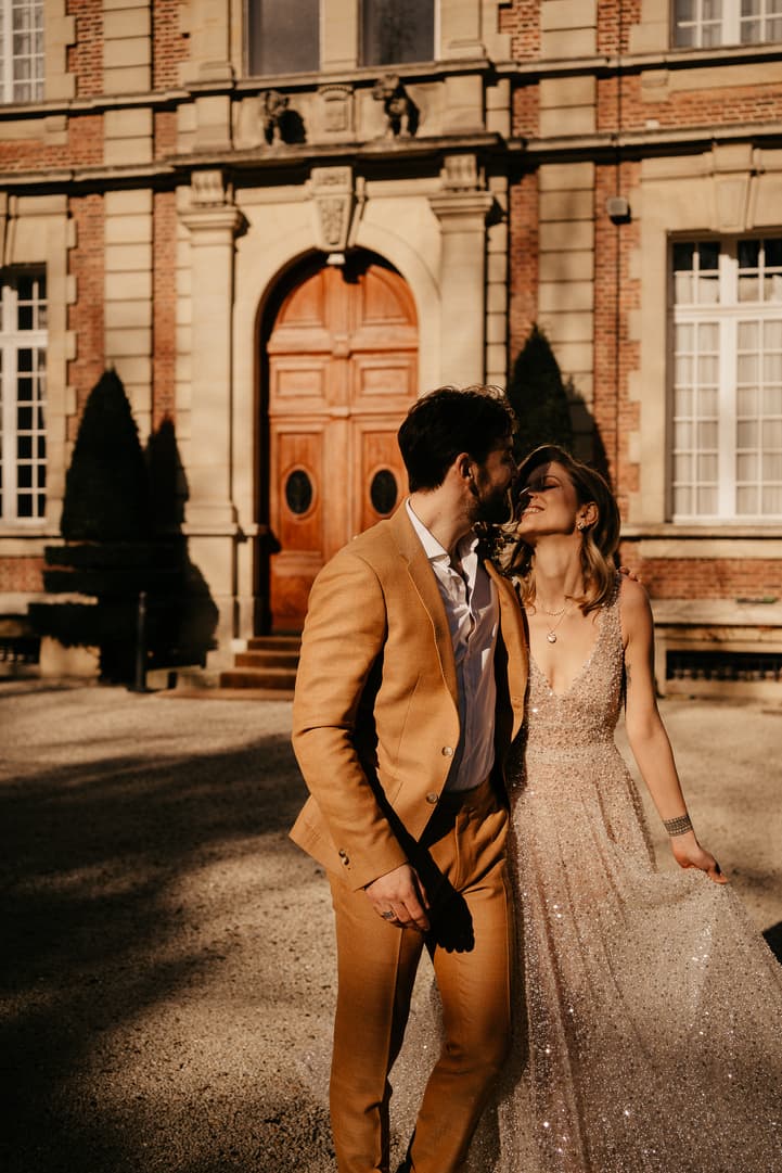 A couple in wedding attire stands on a Paris street before a stately brick mansion in warm sunlight