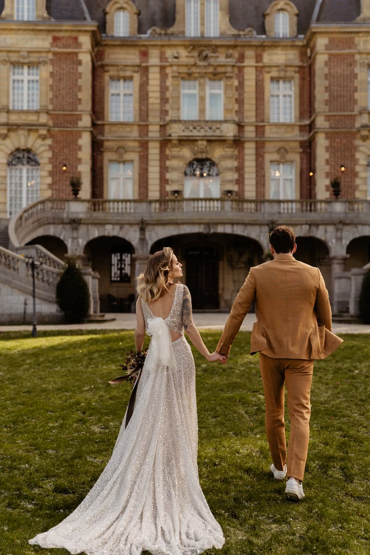 Bride and groom hold hands on a lawn before a grand Paris chateau in warm evening light