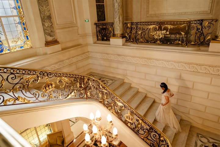 Bride in a flowing gown descends a grand hotel staircase with gold railings and warm light