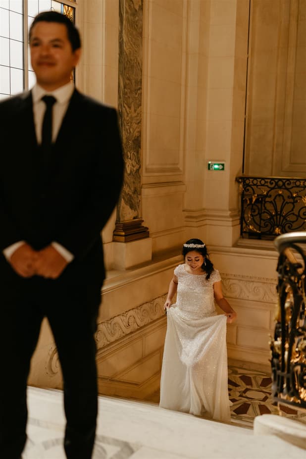 Blurred groom in foreground watches a bride ascending an ornate staircase in warm indoor light