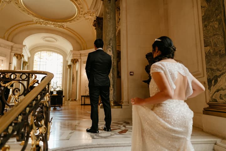 Bride and groom stand in a grand Paris hotel lobby, warm light on marble floors and gilded railings