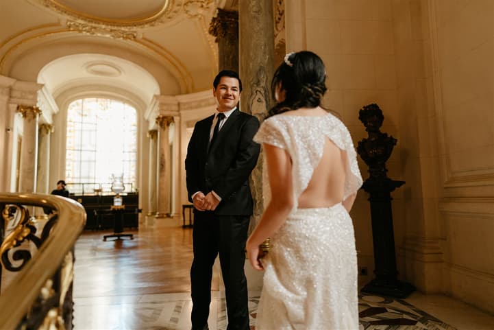 Bride and groom stand in a gilded Paris hall, facing each other in warm window light