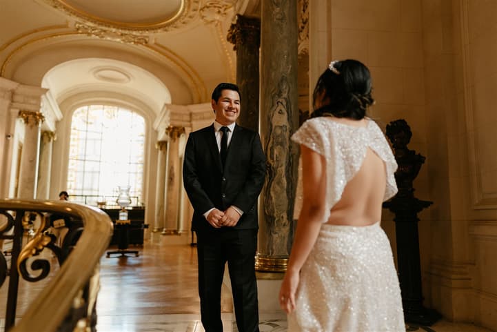 Bride in a beaded backless gown faces smiling groom in a grand Paris hall with warm window light