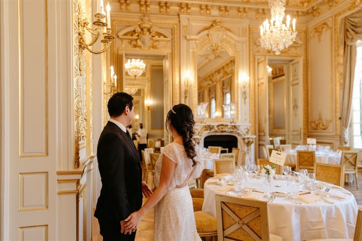 Bride and groom hold hands in an ornate gilded ballroom lit by chandeliers and set tables
