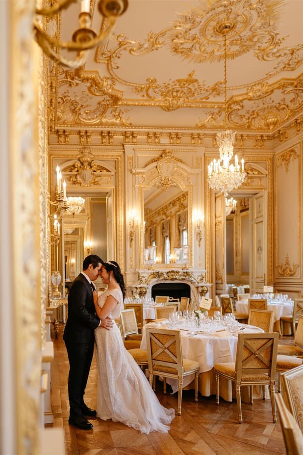Bride and groom embrace in a gilded Paris ballroom with chandeliers and set tables