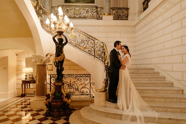 Bride and groom pose on a grand staircase in warm hotel light, beside an ornate black lamp stand