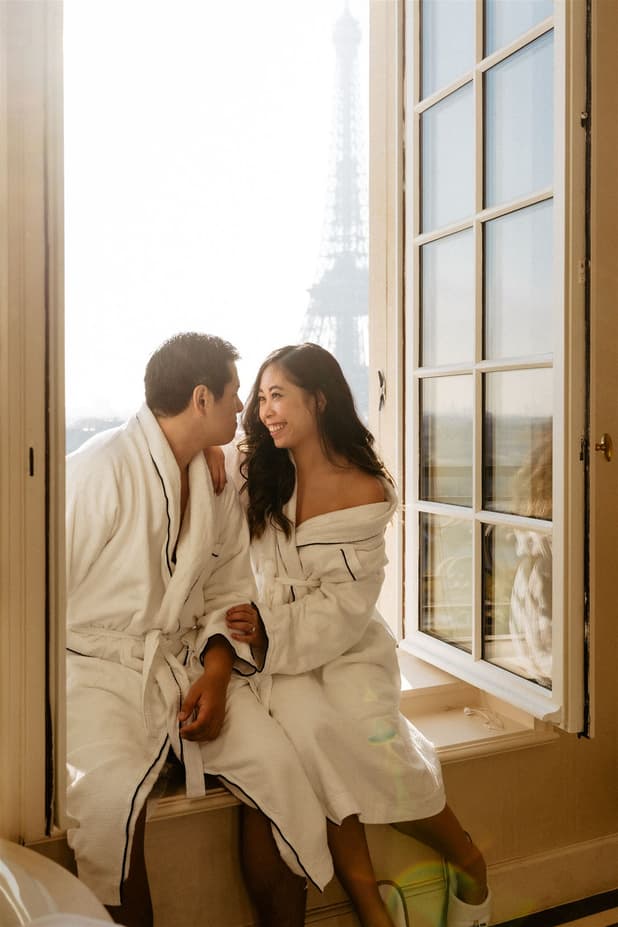 Couple in white robes sit by a Paris window, smiling with the Eiffel Tower softly visible outside