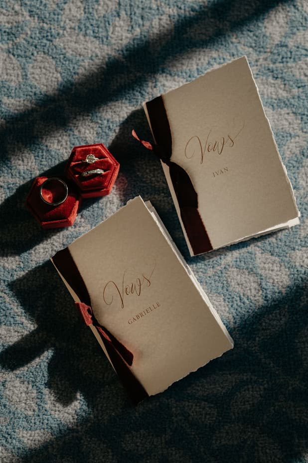 Two red ring boxes and vow books lie on blue fabric in slanted window light