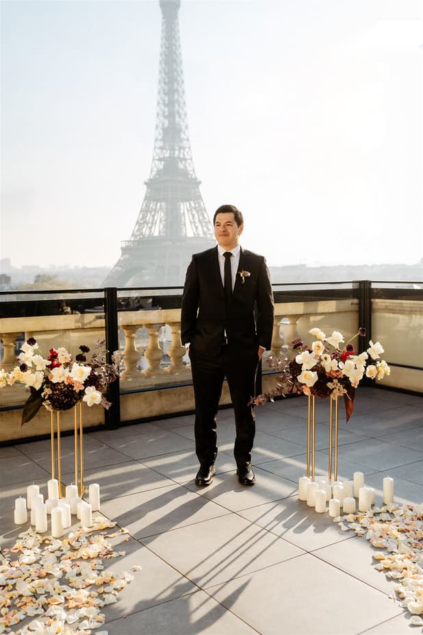 Groom in a black suit stands on a Paris terrace with the Eiffel Tower behind him, candles and flowers below