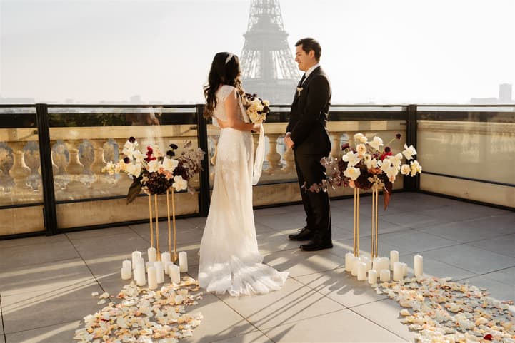 Bride and groom face each other on a terrace with the Eiffel Tower behind, flanked by flowers and candles