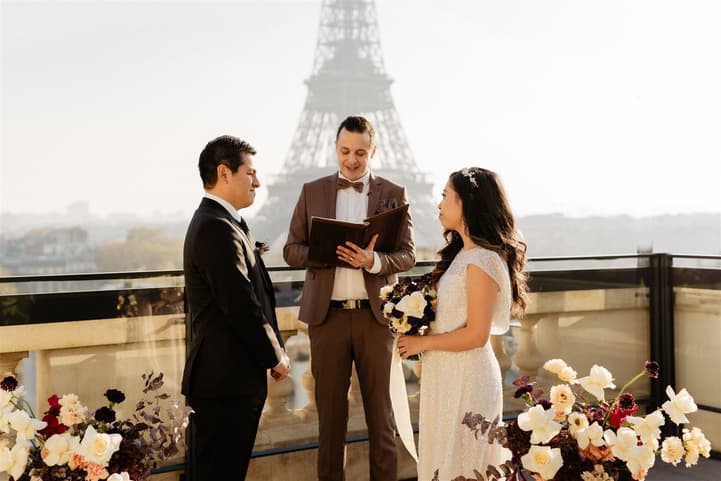 Bride and groom stand with an officiant on a terrace, exchanging vows with the Eiffel Tower behind them