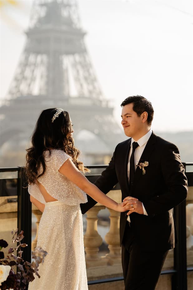Bride and groom hold hands on a terrace in warm light, with the Eiffel Tower blurred behind them
