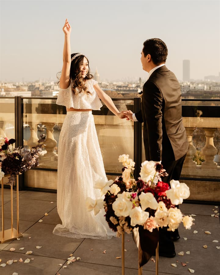 Bride raises one arm while holding groom's hand on a Paris terrace in warm light, with flowers in front