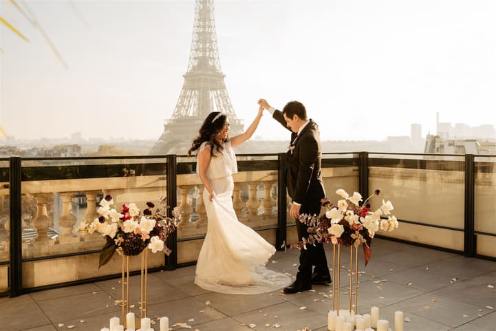 Bride and groom raise joined hands on a terrace with the Eiffel Tower behind, flowers framing them