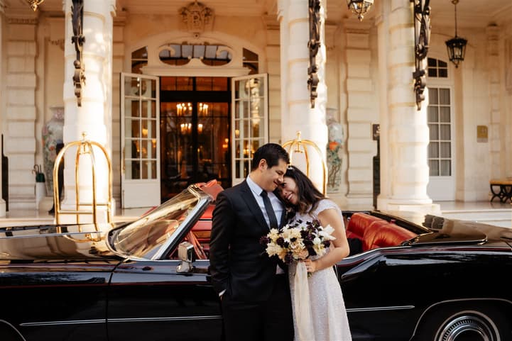 Bride and groom pose beside a black vintage convertible outside a Paris hotel entrance in warm light