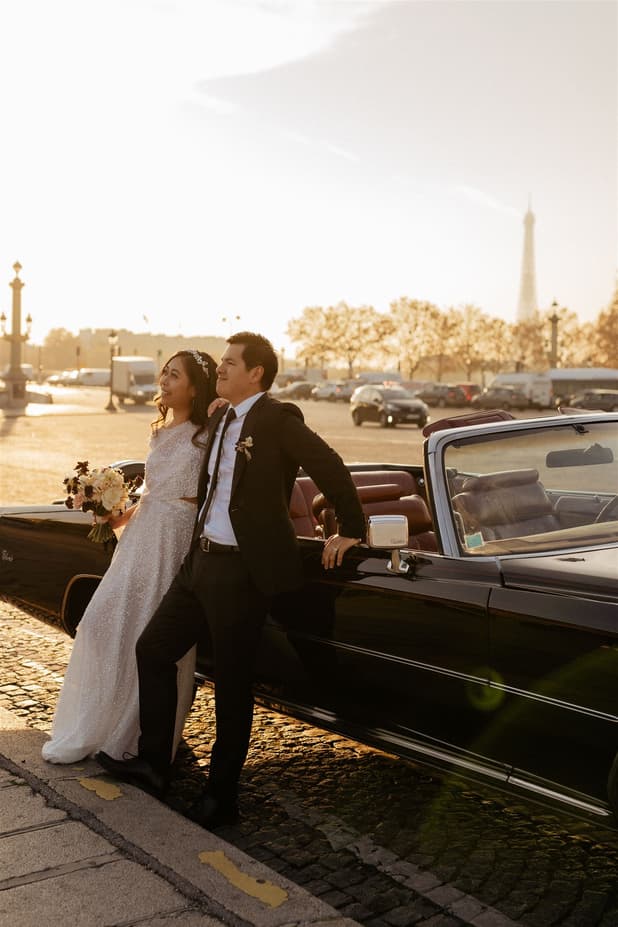 Bride and groom lean by a vintage car on a Paris street, with the Eiffel Tower in soft sunset light