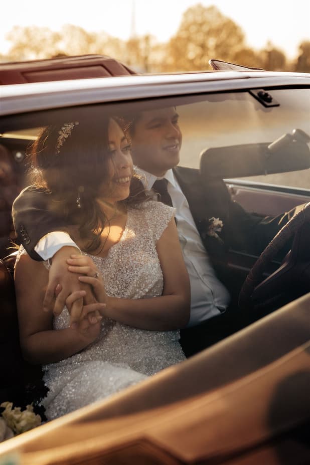 Bride and groom sit in a convertible on a Paris street, holding hands in warm golden light