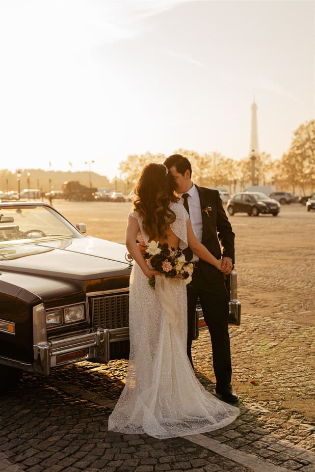 A bride and groom hold hands beside a vintage car on a Paris street at sunset, with the Eiffel Tower behind them