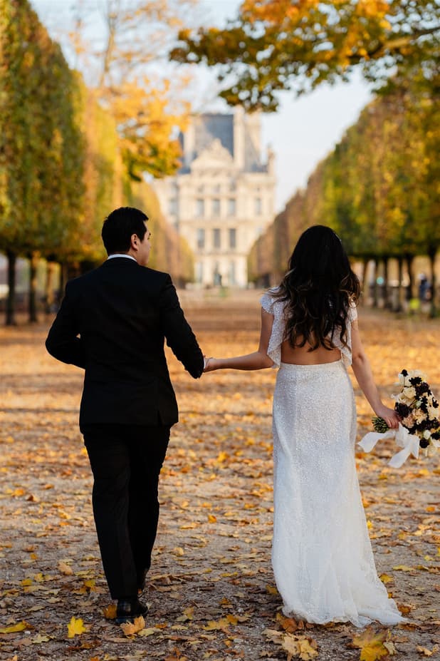 Bride and groom walk hand in hand down a leaf-strewn Paris garden path in warm autumn light
