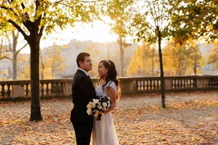 Bride and groom stand in a Paris garden at golden hour, holding a bouquet amid autumn leaves