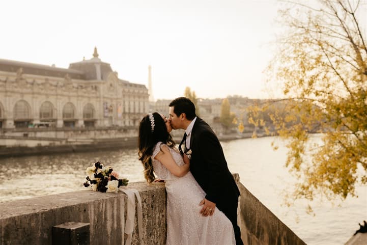 Bride and groom kiss by the Seine at sunset, with a bouquet on the stone wall and golden trees