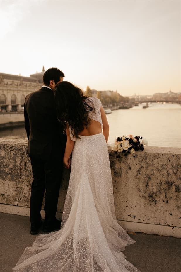 Bride and groom lean over a stone bridge by the Seine at golden hour, with a bouquet on the parapet