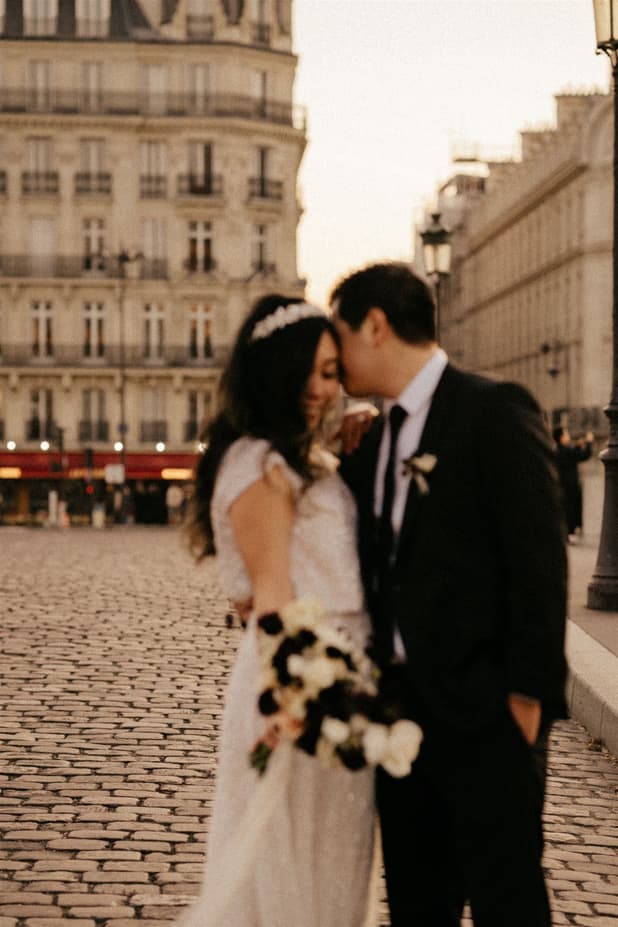 Bride and groom kiss on a Paris street at golden hour, bouquet in hand and cobblestones below