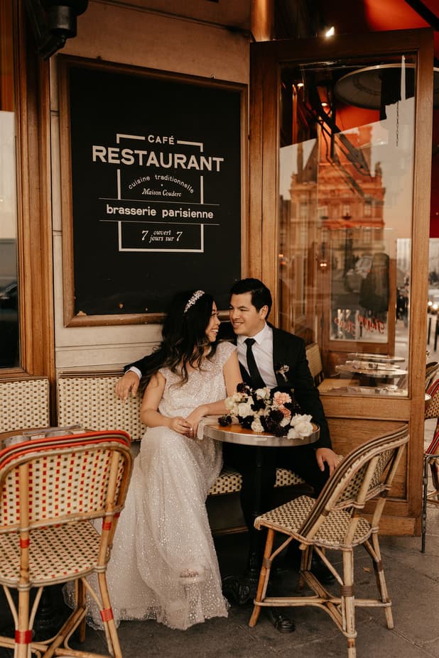 Bride and groom sit at a Paris street café table in warm light, with a bouquet and wicker chairs