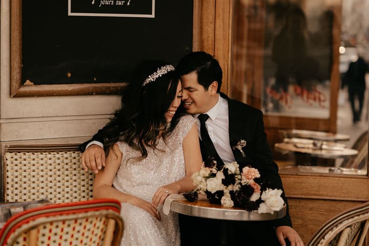 Bride and groom lean together at a Paris café table, with a bouquet and warm window light