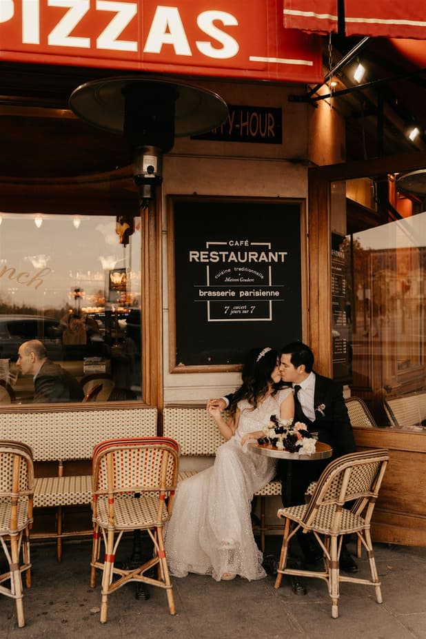 Bride and groom kiss at a Paris café terrace, warm light glowing on wicker chairs and flowers
