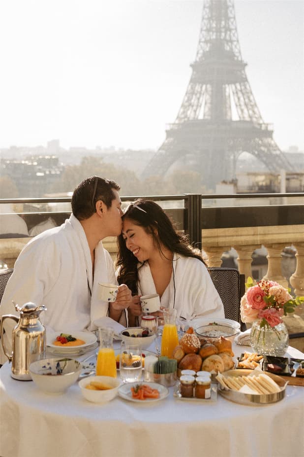 Bride and groom share breakfast on a terrace with the Eiffel Tower behind them and a table of pastries