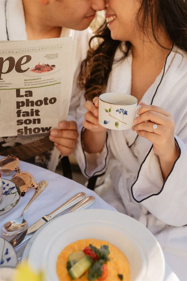 A couple in white robes share breakfast at a table, one reading a magazine and the other holding a floral cup, with warm light and a ring visible