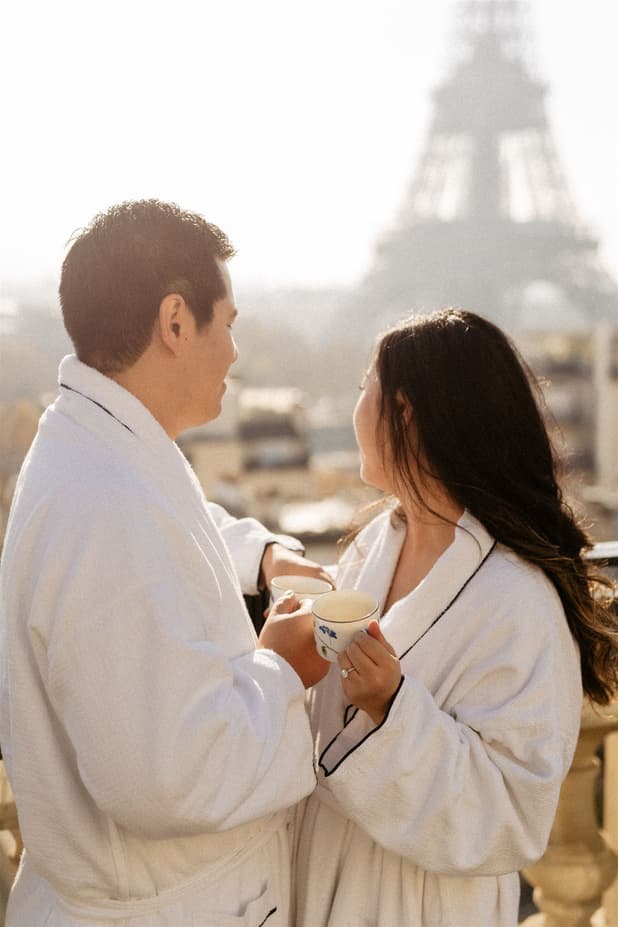 Couple in white robes share tea on a Paris balcony with the Eiffel Tower glowing behind them