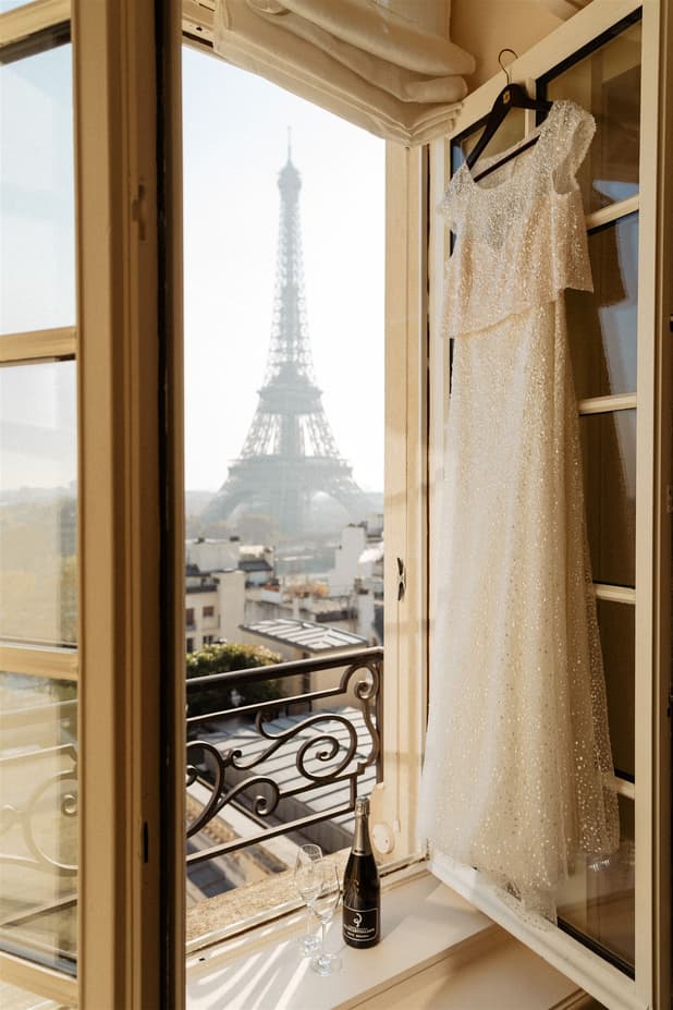 A sparkling wedding dress hangs by a balcony window with the Eiffel Tower beyond in soft morning light