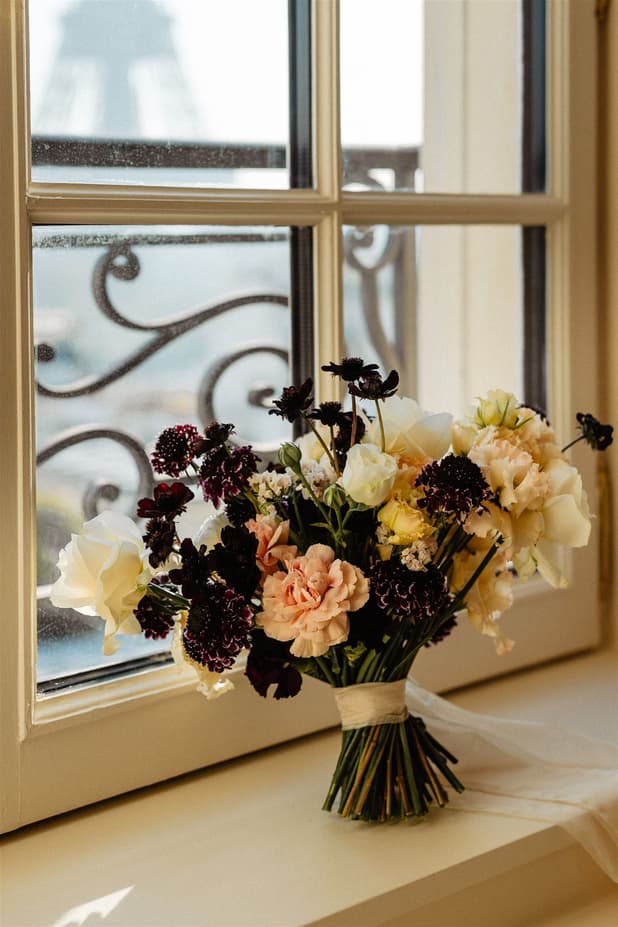 Bouquet of pastel and dark flowers sits on a Paris windowsill in soft daylight with the Eiffel Tower blurred behind it