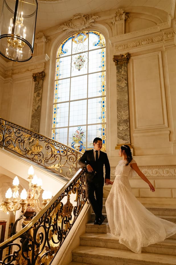 Bride and groom pose on an ornate staircase beneath a stained-glass window in warm golden light