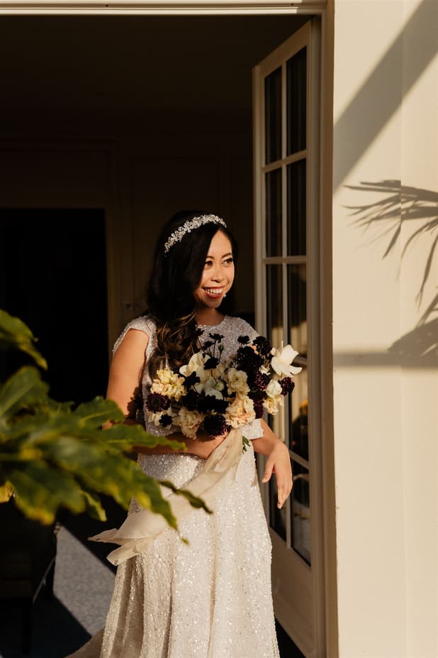 Bride in a sparkling gown holds a bouquet in a sunlit doorway, with plant shadows on the Paris wall