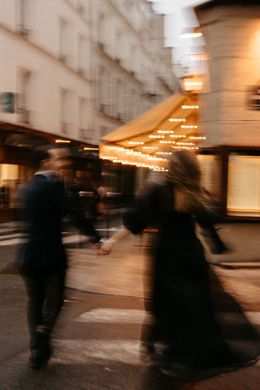 Bride and groom walk hand in hand on a Paris street past a glowing café awning at dusk
