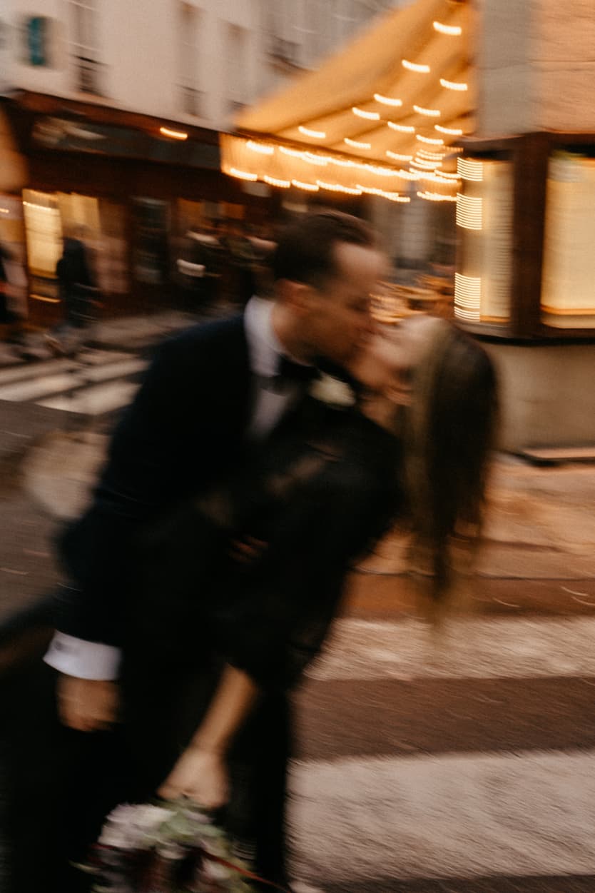 Blurred bride and groom kiss on a Paris street at dusk, with warm café lights and bouquet in hand