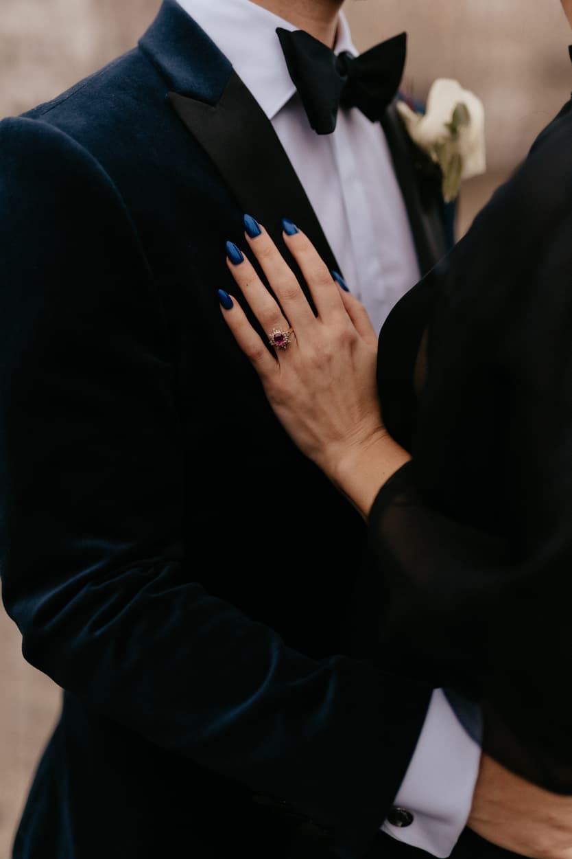 Bride’s blue-nailed hand rests on groom’s velvet tuxedo, showing a pink ring in soft light