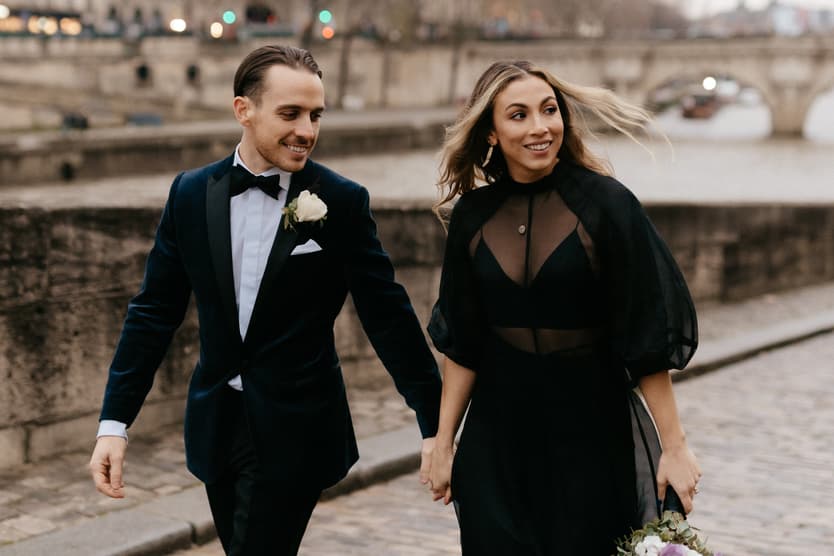 A couple in formalwear walks hand in hand on a Paris street in soft daylight, the bride holding a bouquet