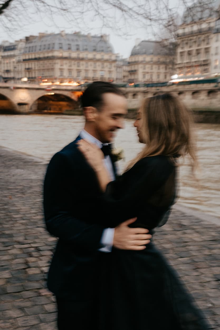 A bride and groom embrace on a Paris riverbank at dusk, with a bridge and glowing buildings behind