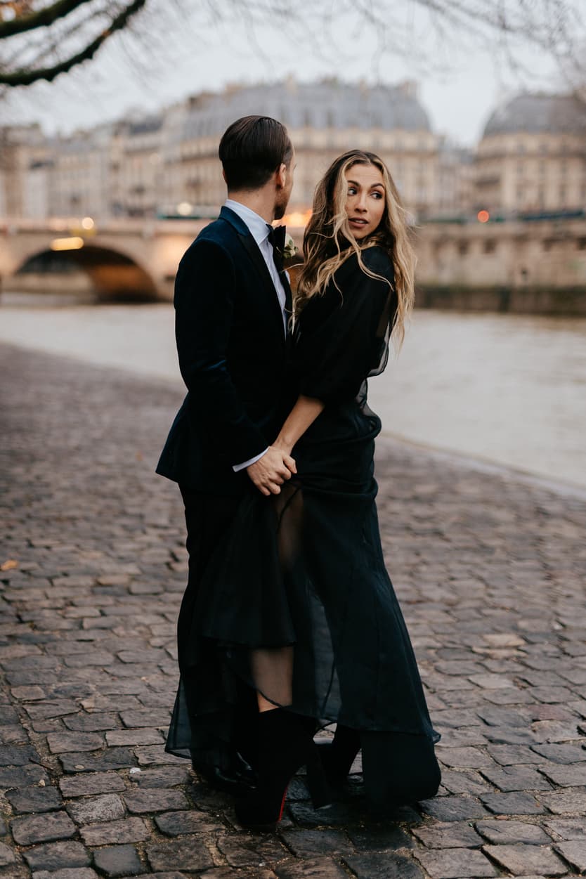 A couple in black holds hands on a cobblestone Paris street by the Seine at dusk