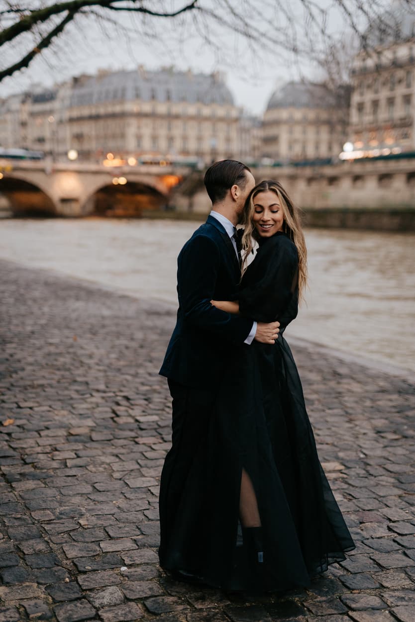 A couple in black embrace on the Seine embankment at dusk, with a bridge and city lights