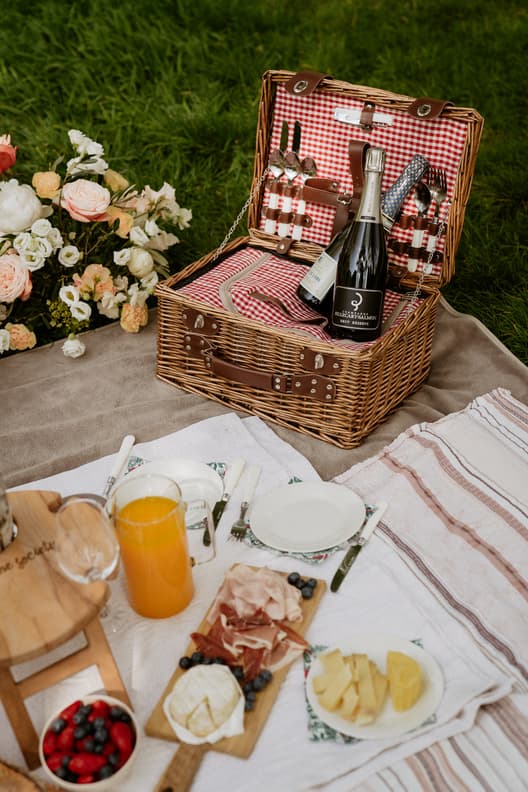 Wicker picnic basket with champagne and cutlery on a blanket beside flowers on green grass