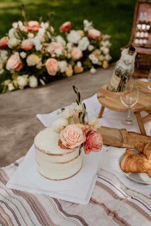A small white wedding cake with peach flowers sits on a picnic table beside croissants, bread and wine in a garden