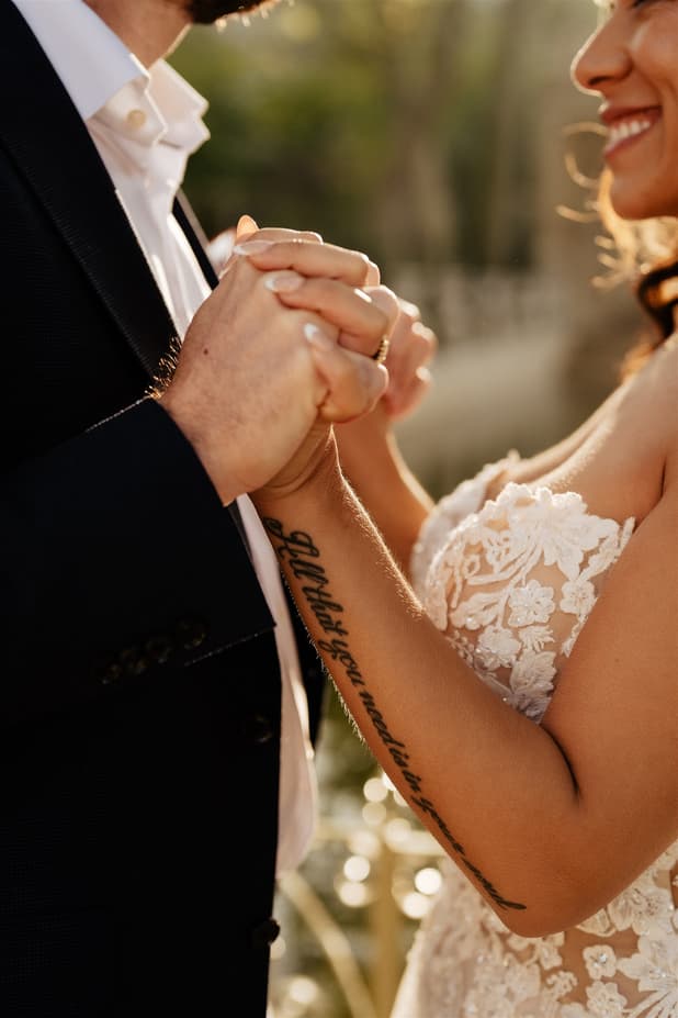 Bride and groom hold hands in warm golden light, her lace dress and forearm tattoo in view