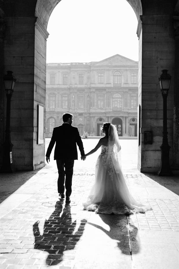 Bride and groom hold hands beneath a Paris archway, backlit on a stone courtyard